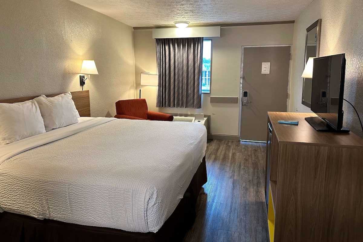 A hotel room with a large bed, a desk with a TV, a chair, a lamp, and a window with curtains.