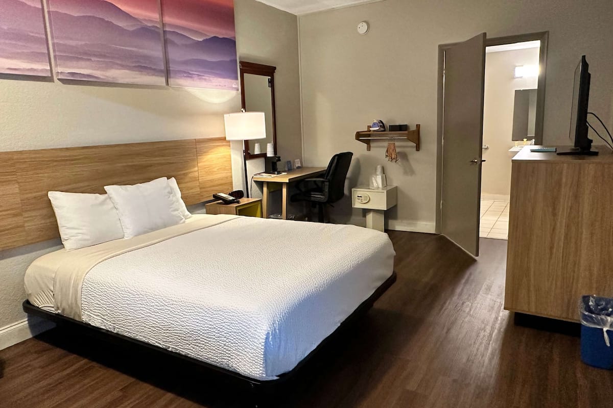 A hotel room with a bed, a desk, a chair, a desk lamp, a mirror, a shelf with toiletries, and a television.