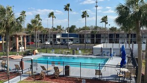 Una piscina con zona de terraza amurallada, rodeada de palmeras y un edificio con varios balcones.
