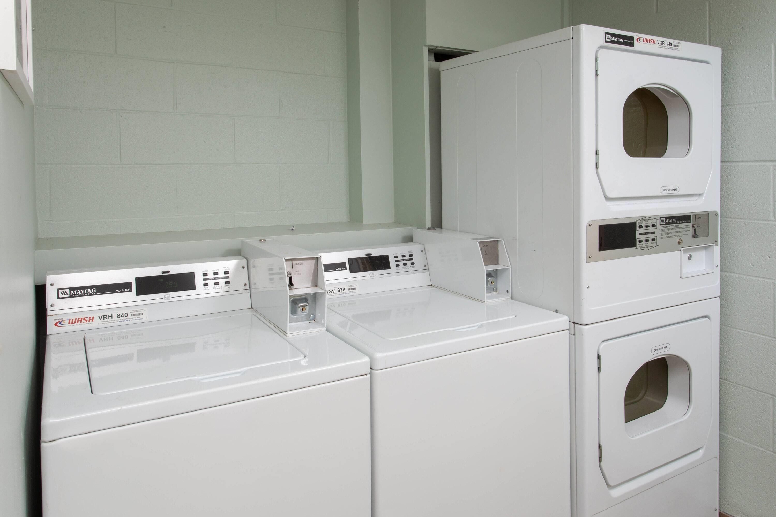 Une buanderie équipée de trois machines à laver et de trois sèche-linge superposés, un mur en briques blanches et une porte donnant sur une autre pièce.