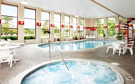An indoor swimming pool with lounge chairs and a hot tub, surrounded by large windows and indoor plants.