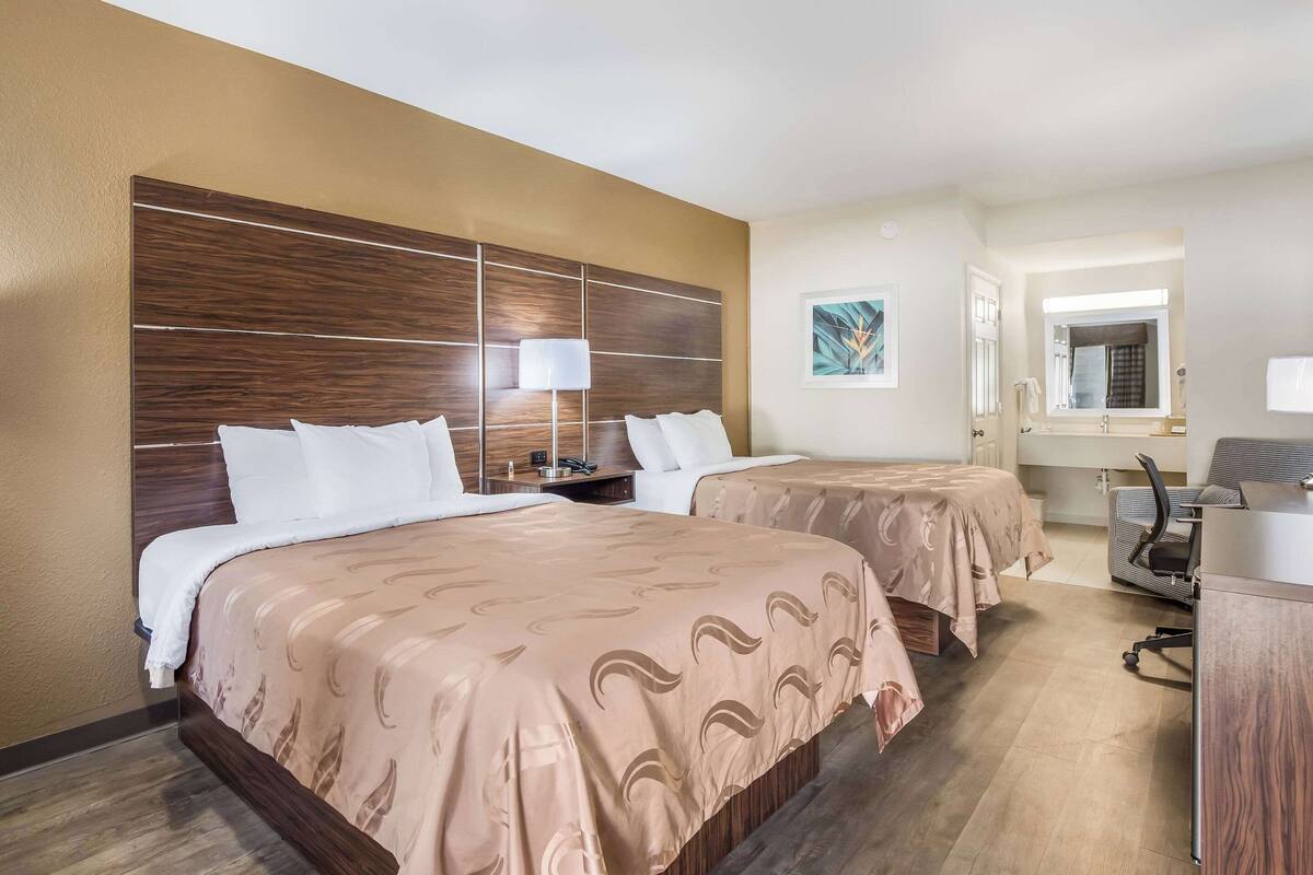 A hotel room with two beds, a desk, a chair, a lamp, and a bathroom visible through an open door.