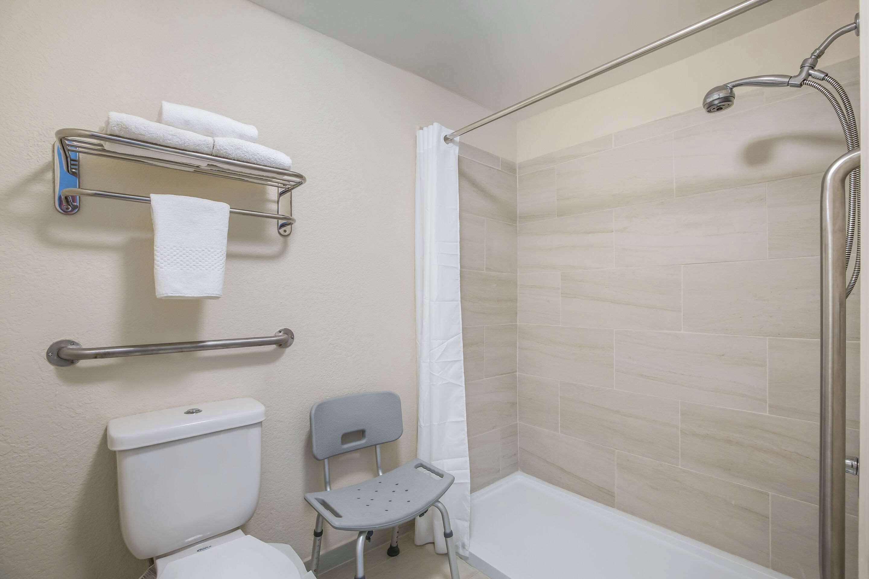 A bathroom with a shower, toilet, grab bars, a wheelchair-accessible bench, and a towel rack.