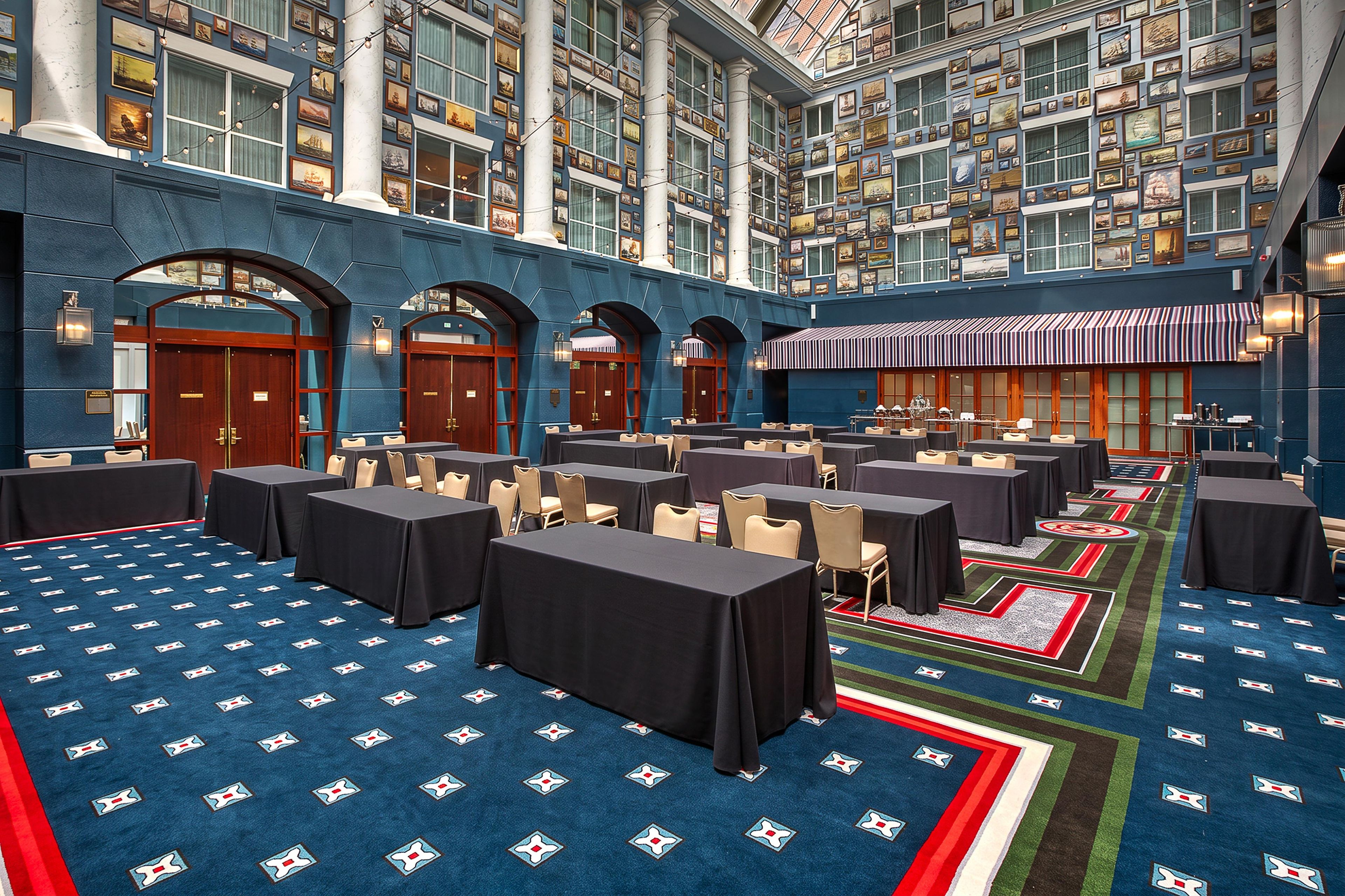 Un hall d’hôtel doté de nombreuses tables et chaises, d’un tapis à motifs et d’une vaste collection de tableaux encadrés accrochés aux murs.