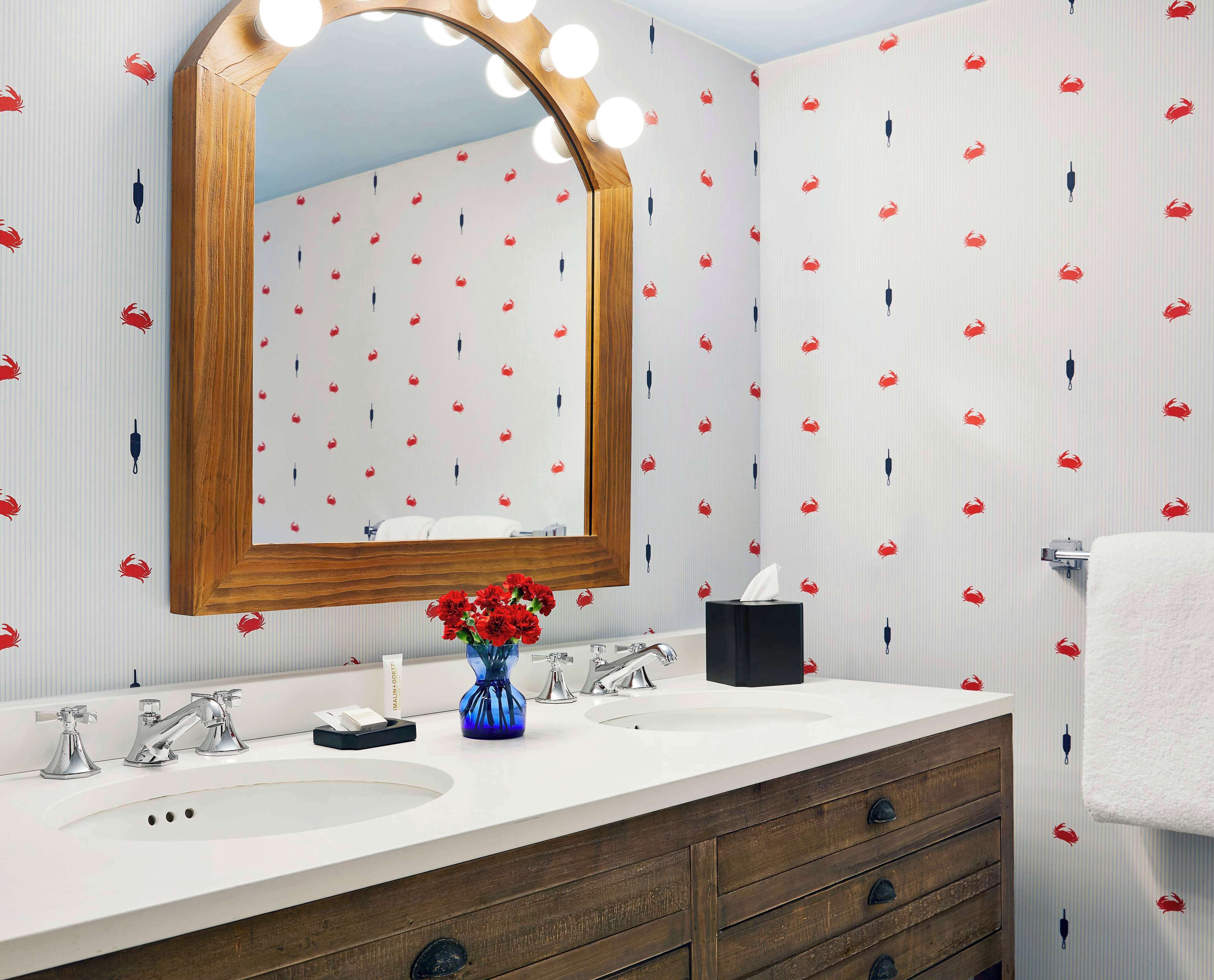 A bathroom with a vanity, two sinks, a mirror with lights, and a patterned wallpaper.