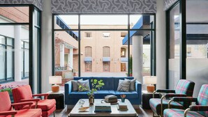 Un área de descanso moderna con un sofá azul, sillas rojas y sillones a cuadros. Una mesa de centro con libros y un jarrón de flores está en el centro. Ventanales grandes ofrecen vistas a un edificio de ladrillo con toldos.