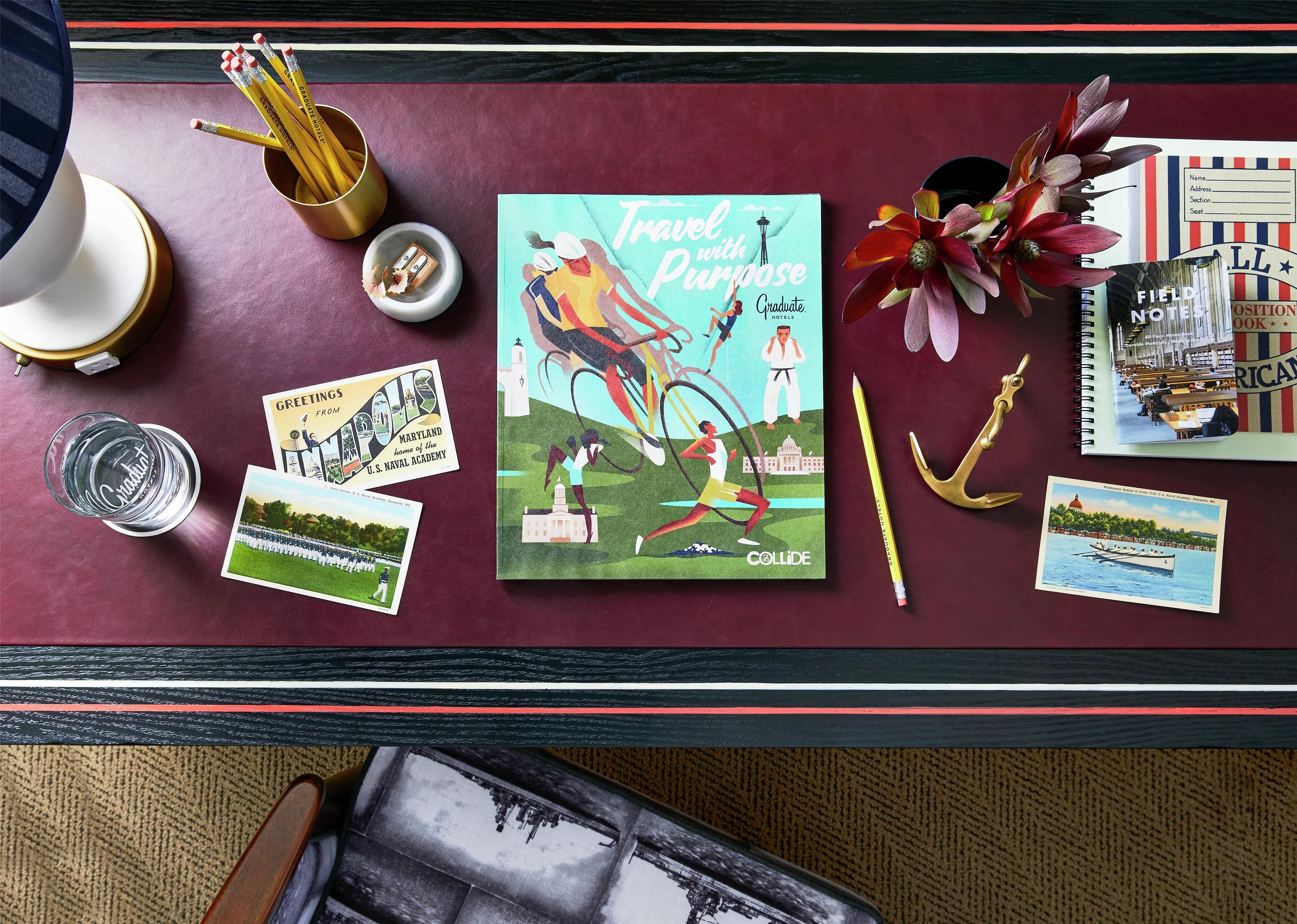 A desk with a book titled 'Travel with Purpose', a notebook, a pencil, a pair of scissors, a glass of water, and a decorative flower.