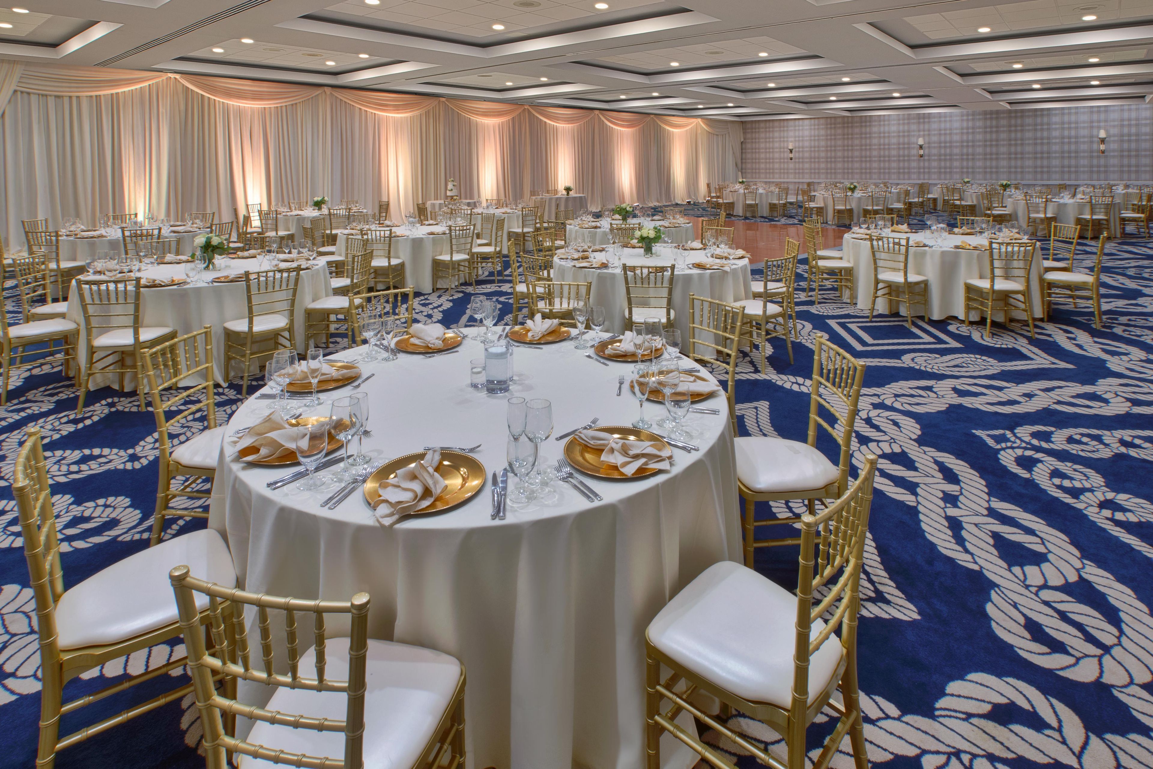 A banquet hall with round tables, gold chairs, and white tablecloths.