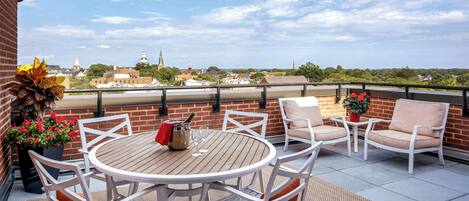 A rooftop terrace with a round wooden table, white chairs, and a view of a cityscape.