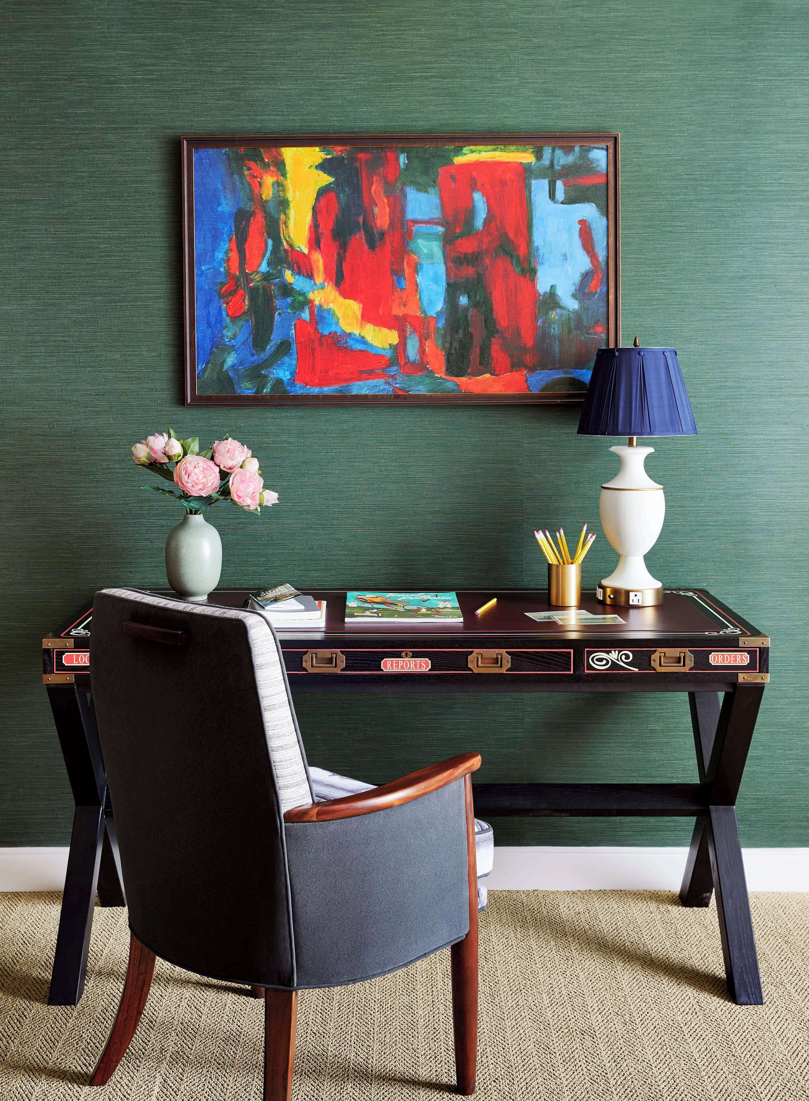 A desk with a vase of flowers, a lamp, and a colorful abstract painting on the wall.