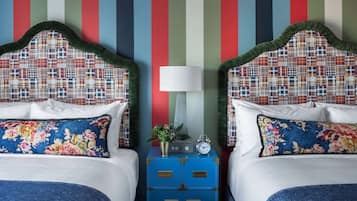 Une chambre d’hôtel avec deux lits, chacun doté d’une tête de lit à carreaux, de draps blancs et d’oreillers à motifs floraux. On y trouve une commode bleue avec une horloge et une petite plante sur la table de chevet.