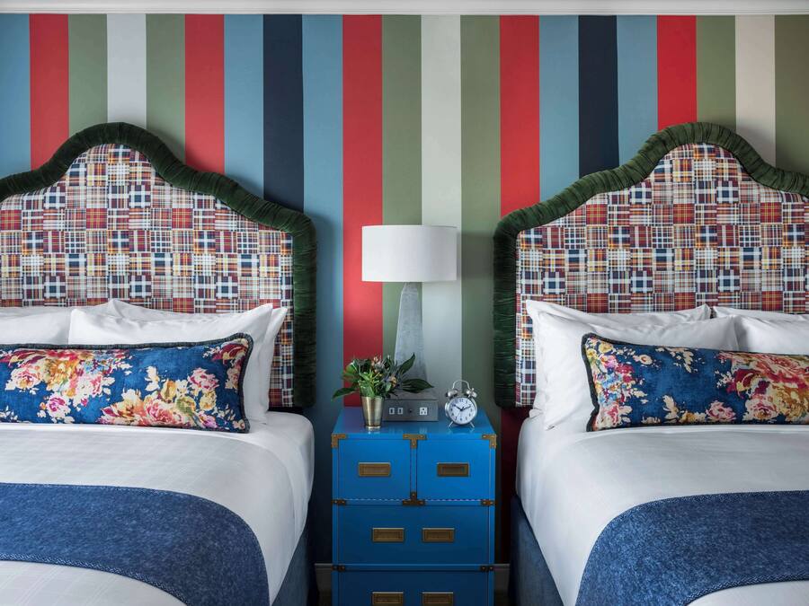 Une chambre d’hôtel avec deux lits, chacun doté d’une tête de lit à carreaux, de draps blancs et d’oreillers à motifs floraux. On y trouve une commode bleue avec une horloge et une petite plante sur la table de chevet.