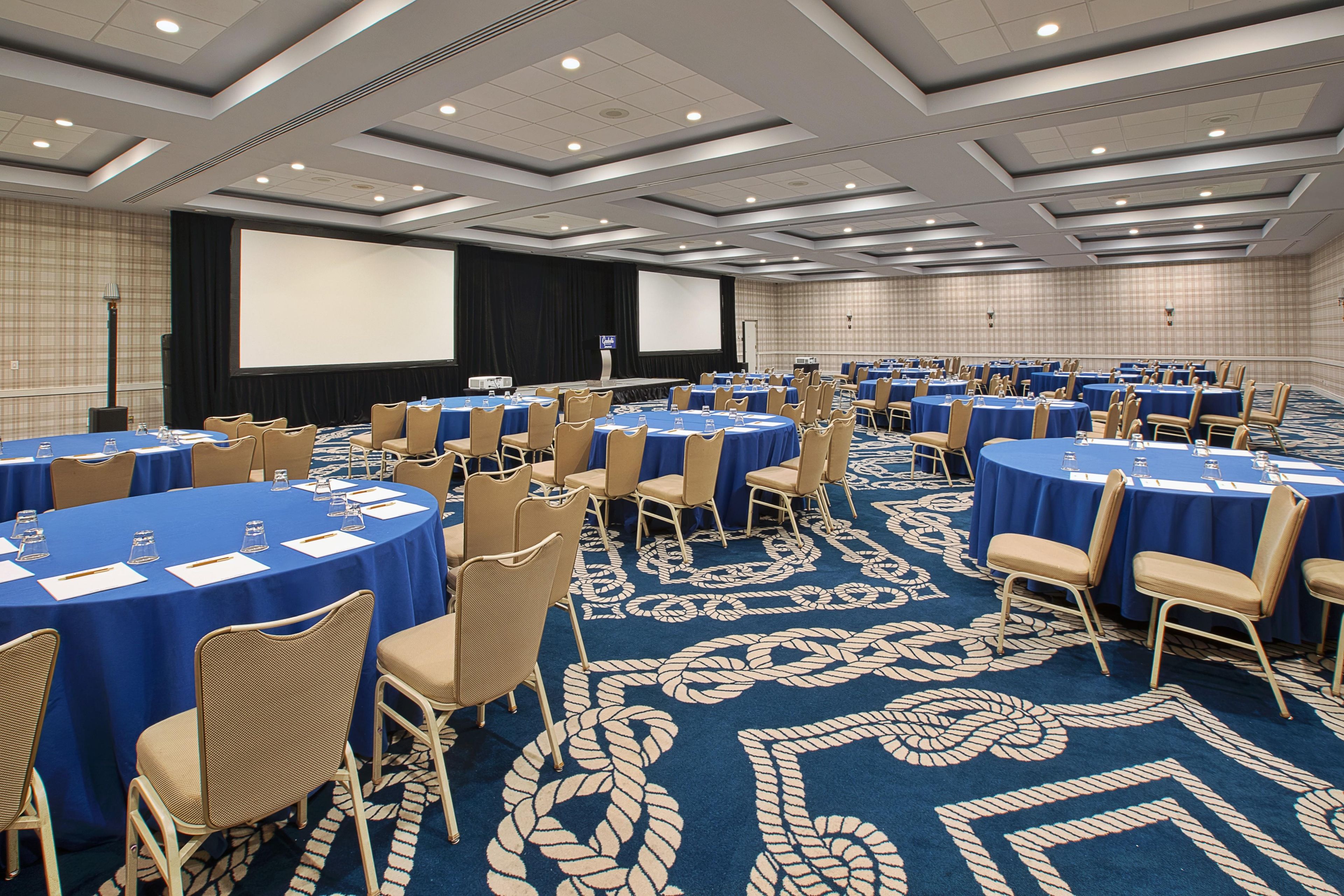 Sala de conferências com mesas redondas, cadeiras, tela de projetor e piso coberto por carpete.