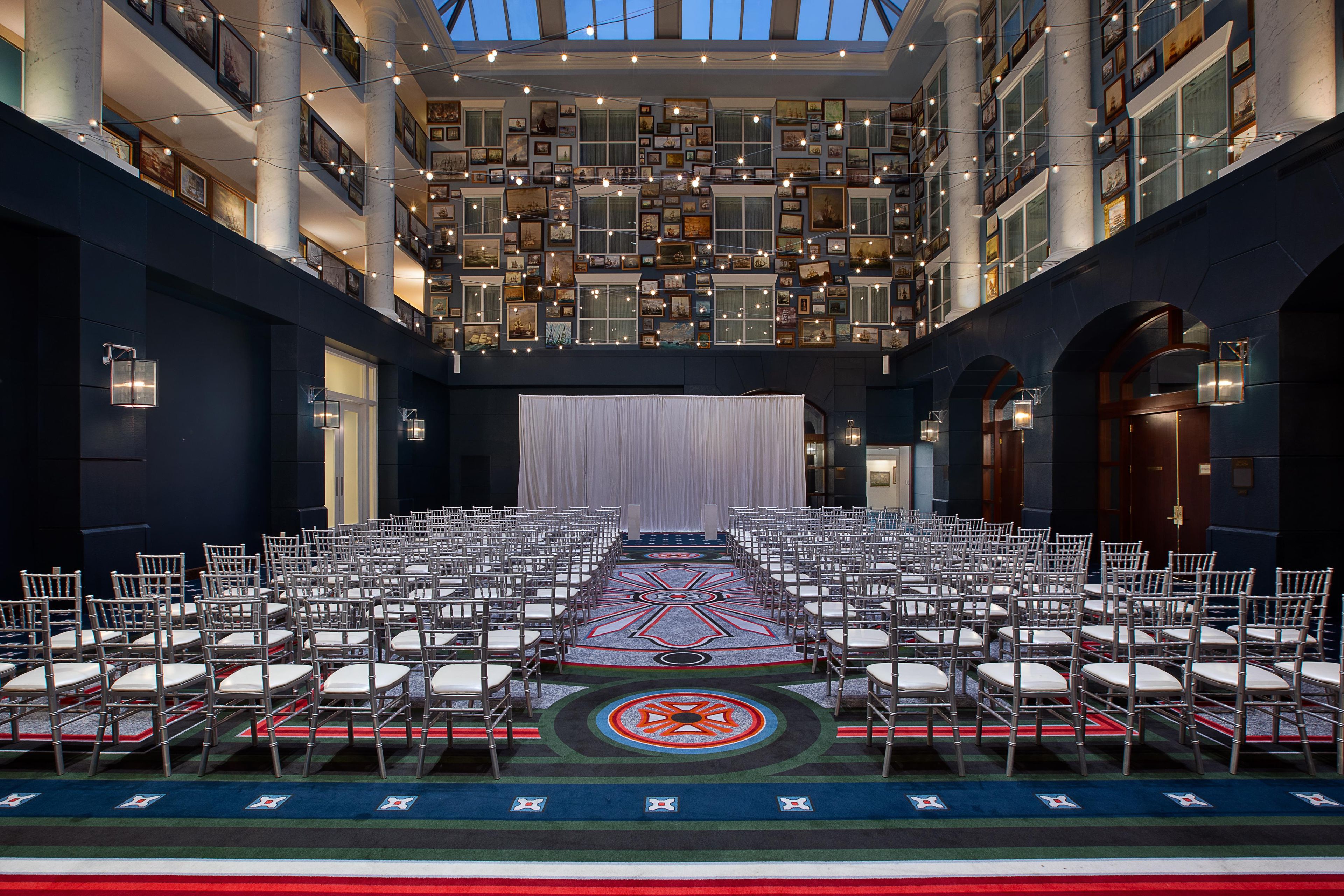 A spacious hall with rows of chairs arranged for an event, a patterned carpet, and a backdrop of framed pictures.
