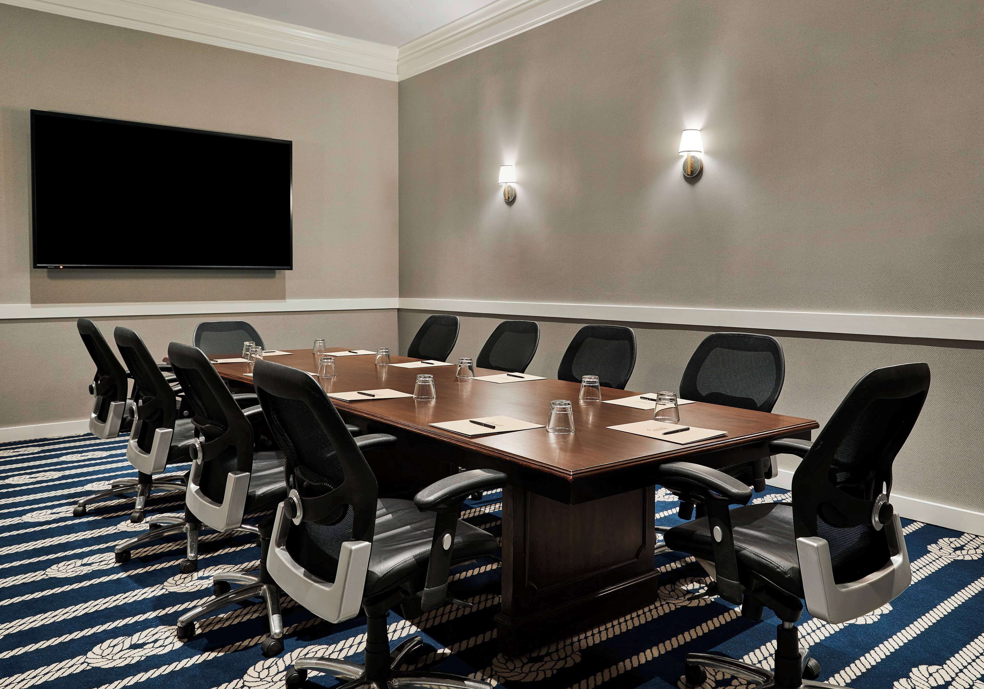 Un salón de conferencias con una mesa grande, sillas negras, un televisor de pantalla plana y dos luces fijadas a la pared.