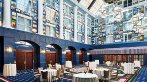 Un vestíbulo de hotel con techo alto, amplias ventanas y una decoración en tonos azul y blanco. Mesas y sillas dispuestas para comer o reuniones.
