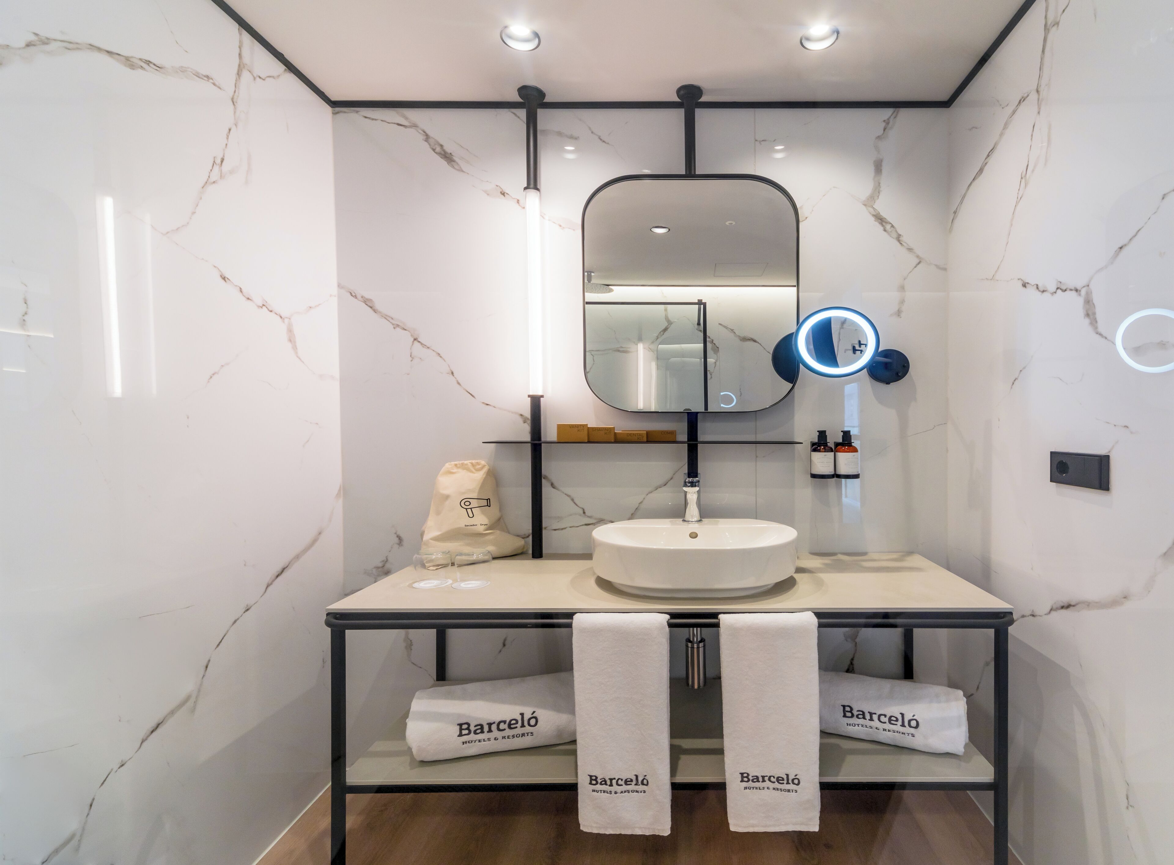 A modern bathroom with a marble wall, a round mirror, a sink, and towels with 'Barceló Hotels & Resorts' branding.