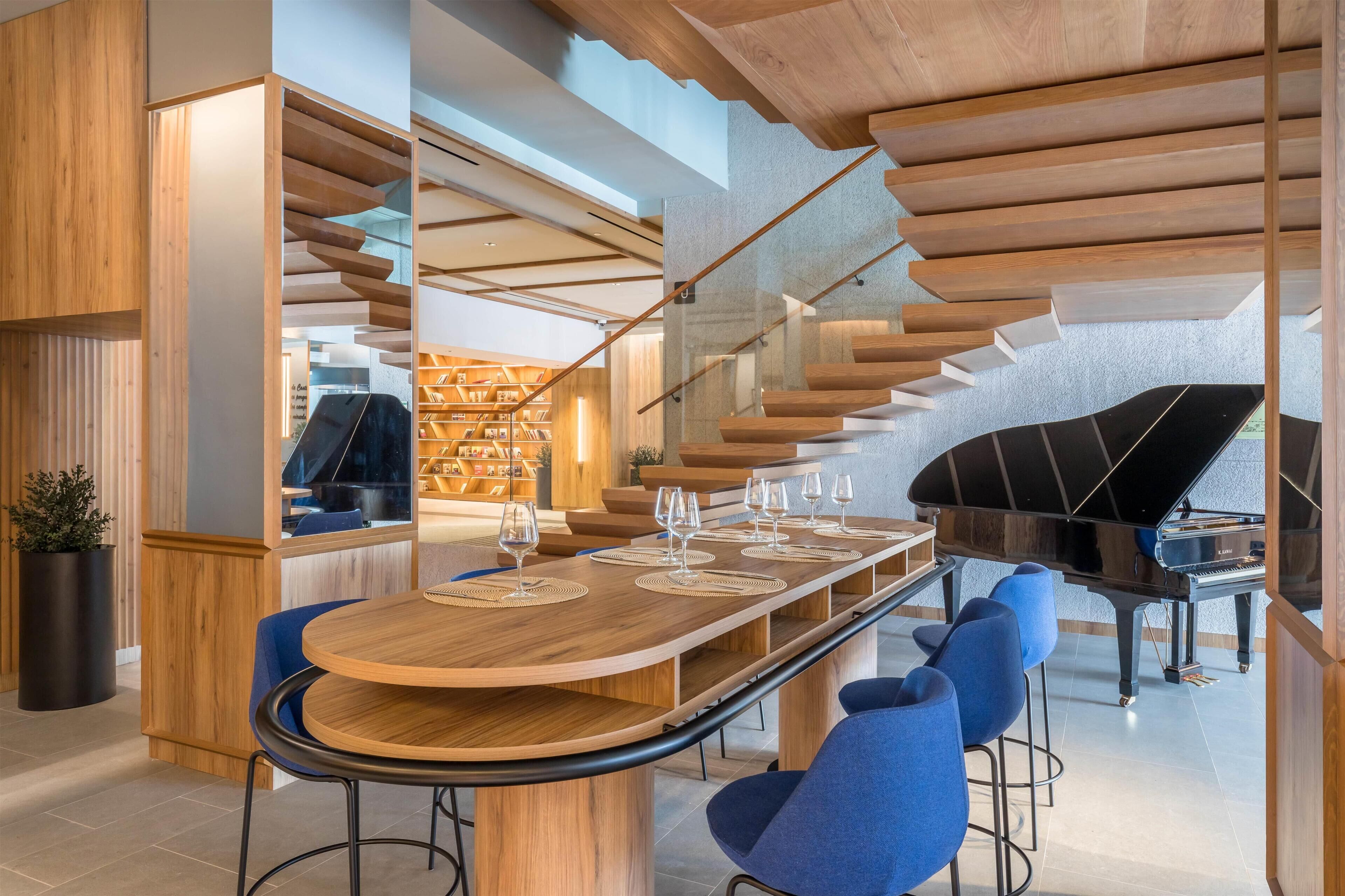 A modern interior with a long wooden table, blue chairs, a grand piano, and a staircase with wooden railings.