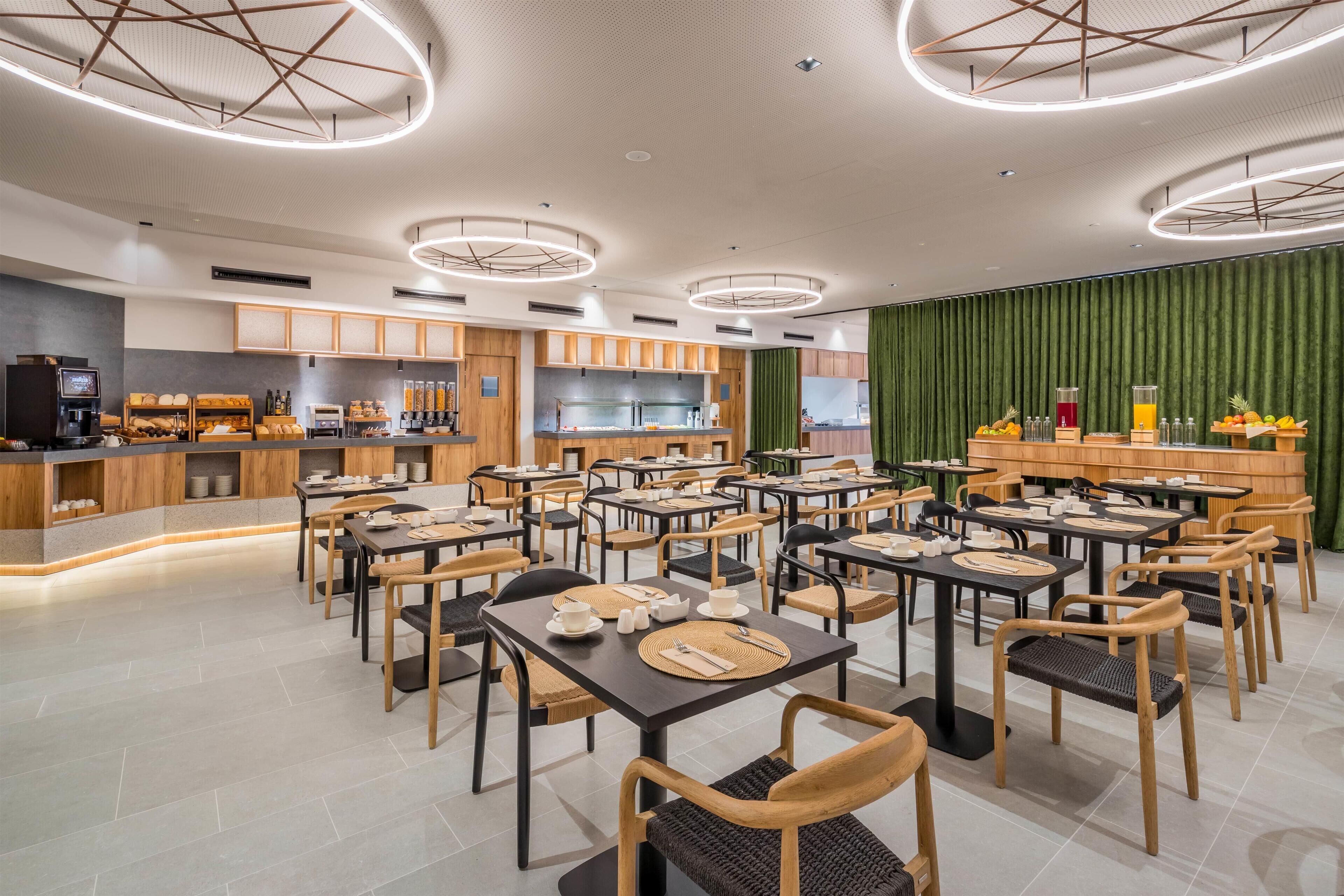 A modern dining area with multiple tables and chairs, a kitchen in the background, and a green curtain wall.