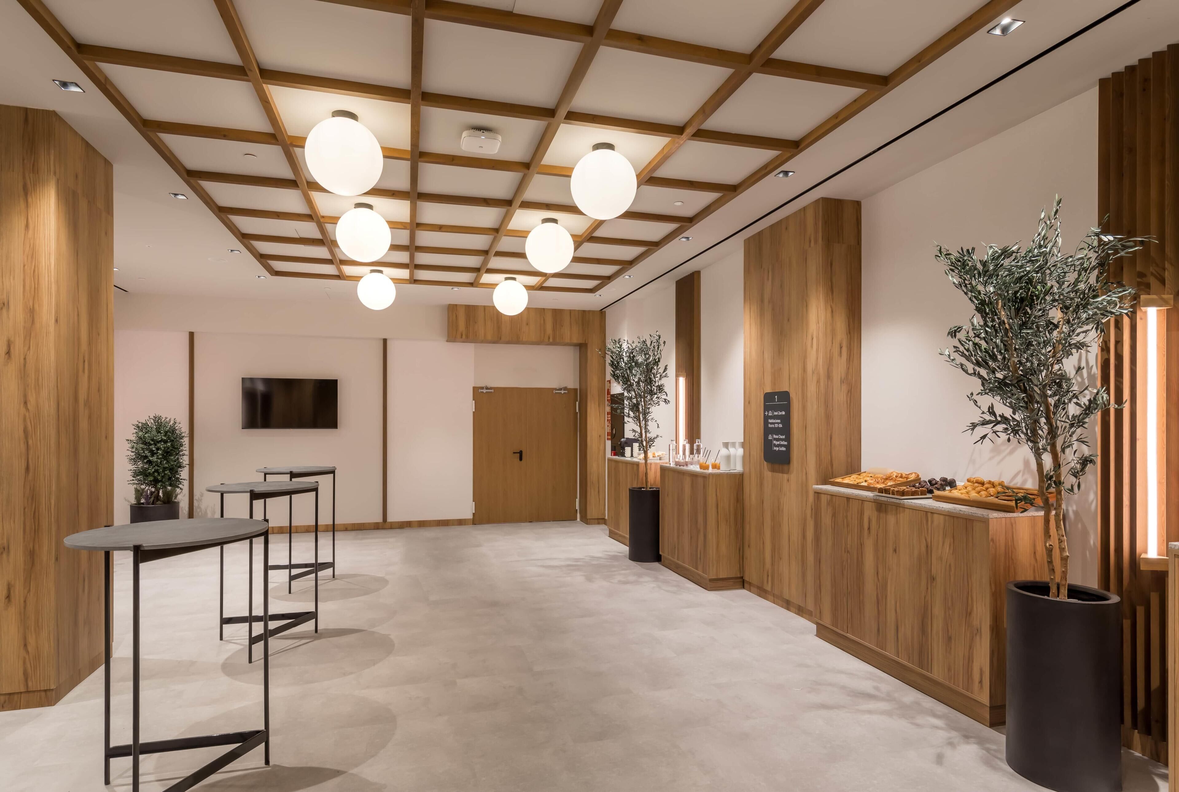 Un vestíbulo moderno con paneles de madera, un mostrador de recepción, un televisor de pantalla plana y un área de recepción con una planta y aperitivos.