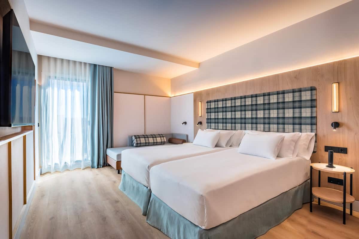 A hotel room with two beds, a TV, a bedside table, and a large window with curtains.