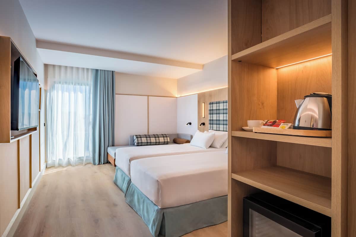 A hotel room with two beds, a television, a coffee maker, and a shelf with a book and a cup.