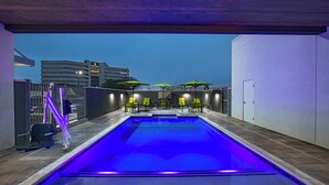 Piscina con iluminación azul, rodeada de sillas y sombrillas, con un paisaje urbano de fondo.