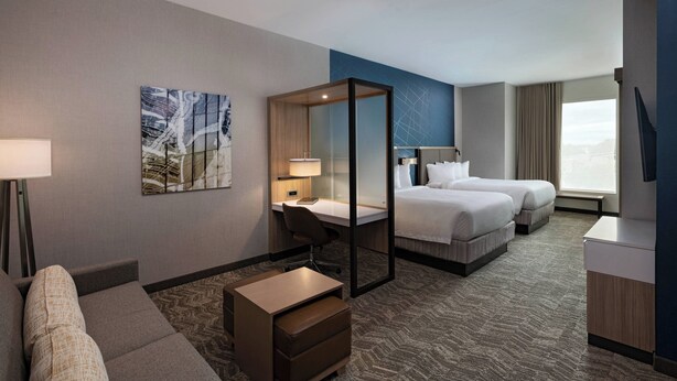 Habitación de hotel con escritorio, dos camas, sofá, mesita, silla y un ventanal grande.