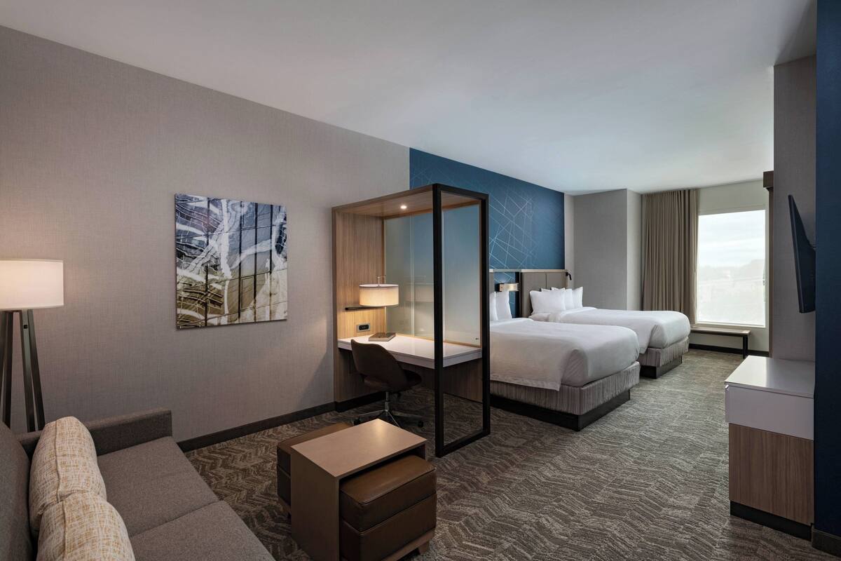 A hotel room with a desk, two beds, a sofa, a small table, a chair, and a large window.