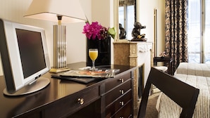 A hotel room with a desk, a computer monitor, a lamp, a vase with flowers, a glass of orange juice, a magazine, a mirror, a bed, and a window with curtains. - Hotel Vaneau Saint Germain (Paris)