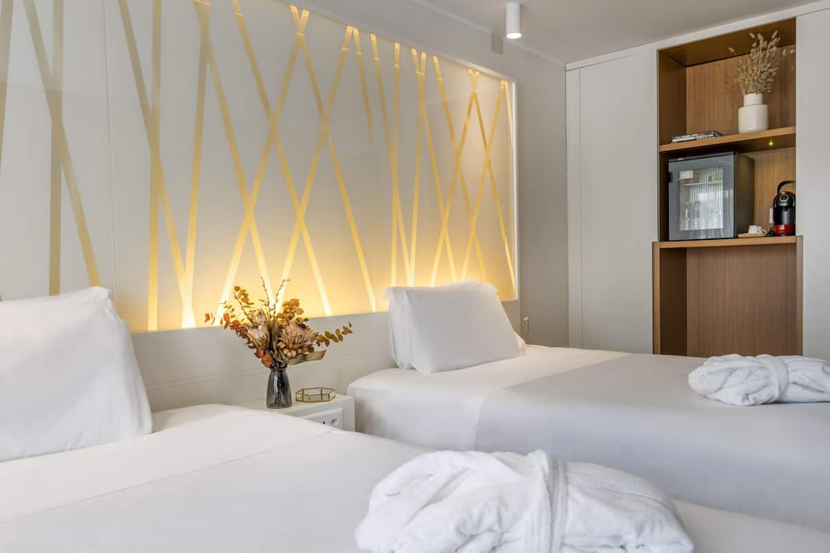 Une chambre d’hôtel avec deux lits, une table de chevet ornée d’un vase de fleurs, une étagère en bois avec un réfrigérateur et une bouilloire, et un mur décoré d’un motif lumineux géométrique.