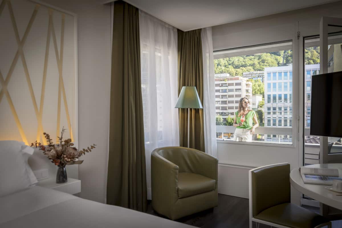 Une chambre d’hôtel avec un lit, une chaise, une table, une lampe et une fenêtre donnant sur des immeubles et des arbres.