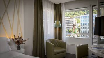 A hotel room with a bed, a chair, a table, a lamp, and a window with a view of buildings and trees.