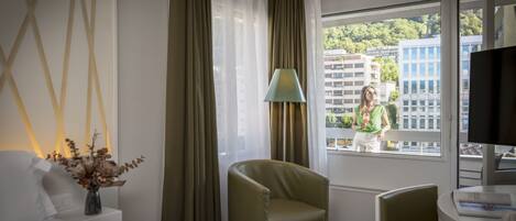 A hotel room with a bed, a chair, a table, a lamp, and a window with a view of buildings and trees.