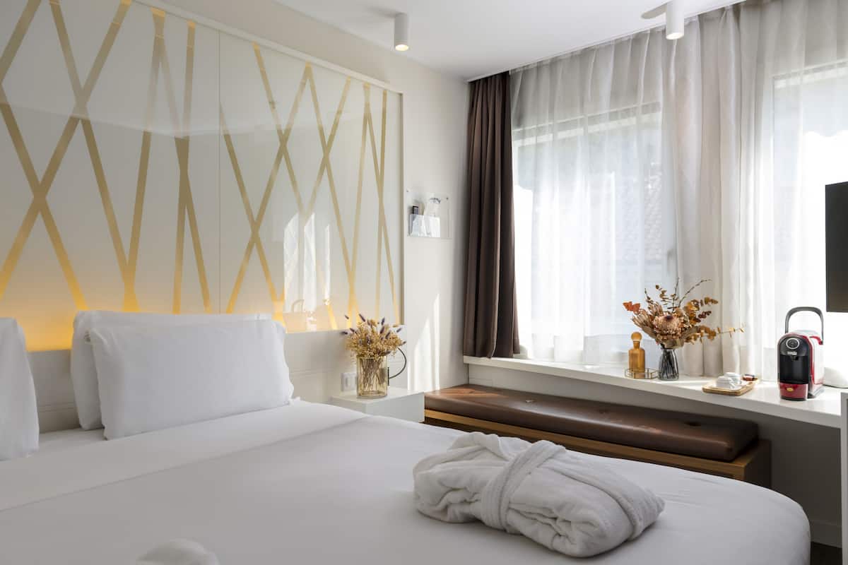 Une chambre d’hôtel moderne avec un grand lit, une table de chevet ornée d’un vase de fleurs, une fenêtre avec des rideaux et une machine à café posée sur la table.