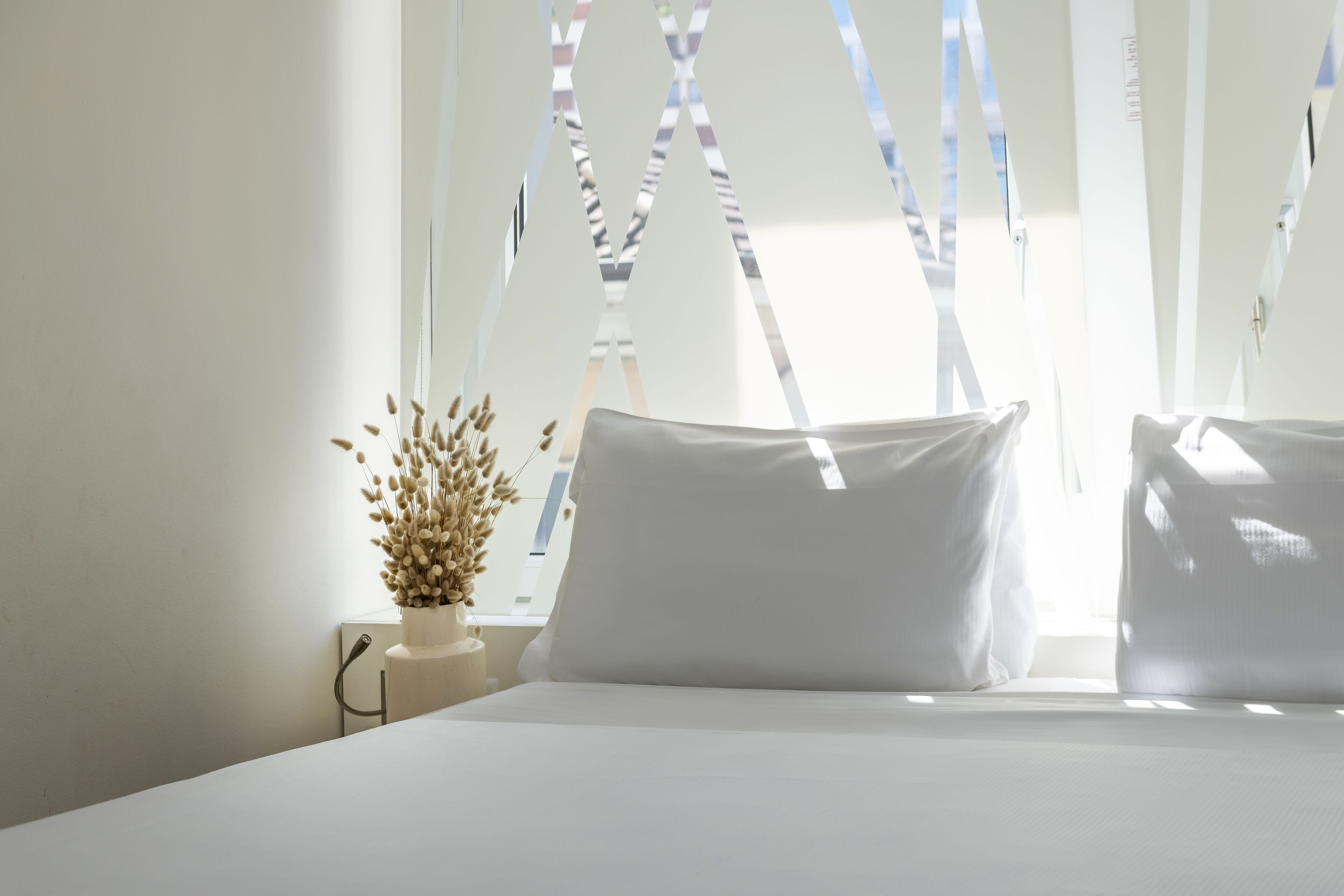 Un lit bien fait, avec du linge de lit blanc, un vase contenant des fleurs séchées et une fenêtre dont les stores sont partiellement baissés.