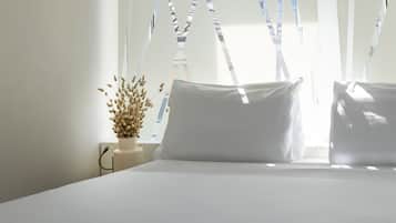 Un lit bien fait, avec du linge de lit blanc, un vase contenant des fleurs séchées et une fenêtre dont les stores sont partiellement baissés.