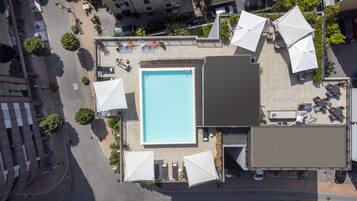Vista aérea de un área de piscina en la azotea, con una piscina rectangular, rodeada de mobiliario exterior y sombrillas.