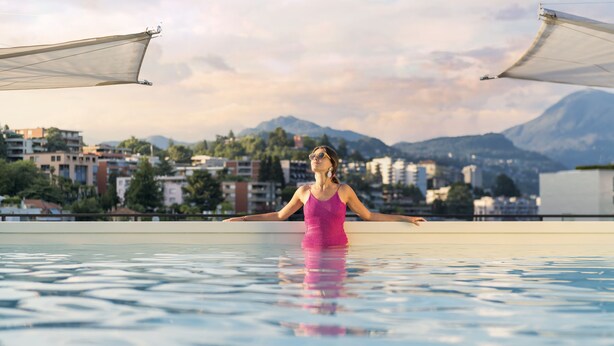 Una persona con traje de baño rosa sentada al borde de una piscina, con un paisaje urbano y montañas de fondo.
