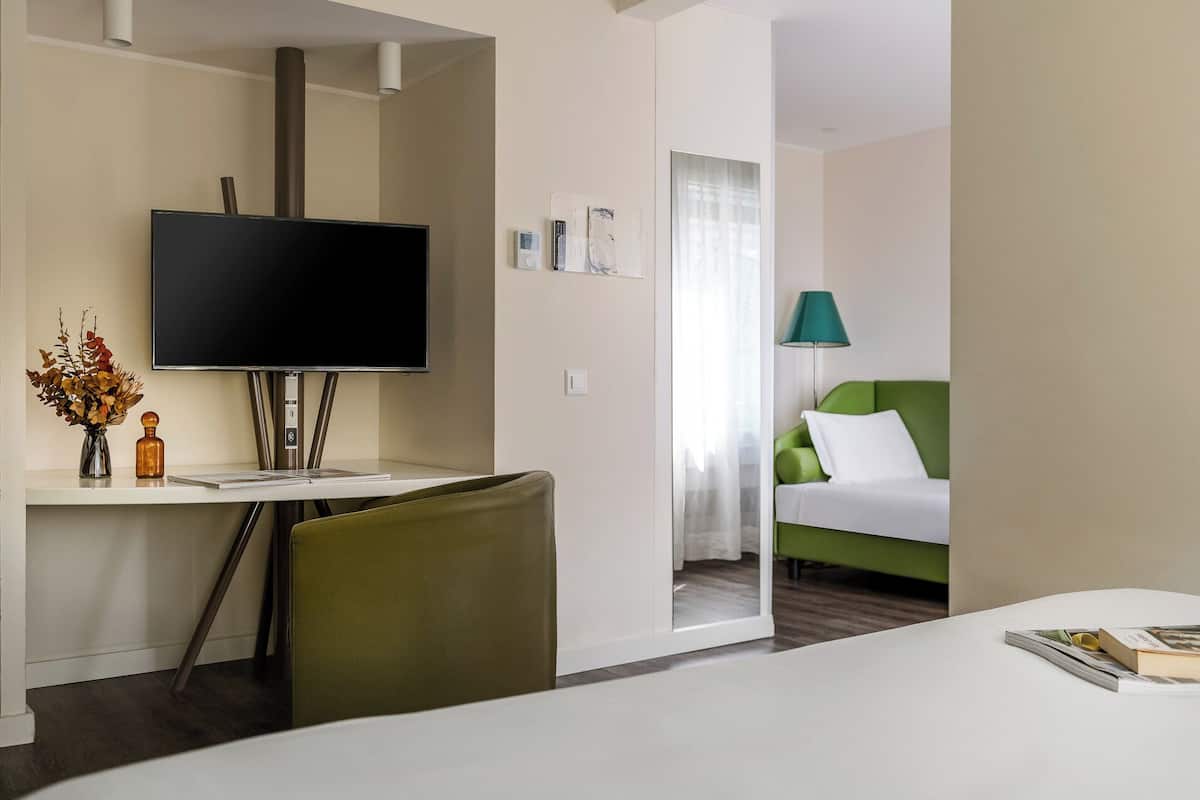 Une chambre d’hôtel équipée d’un téléviseur à écran plat, d’un bureau agrémenté d’un vase et d’une bouteille, d’un fauteuil vert, d’un lit avec une tête de lit verte et d’une lampe.