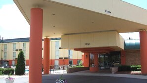 A hotel entrance with a covered area supported by orange columns, a number '3750' above the entrance, and potted plants on the sidewalk. - A Victory Inn & Suites - Ann Arbor (Ann Arbor)