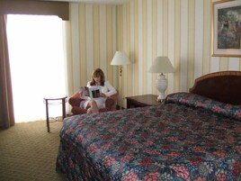 Una habitación de hotel con cama, una silla, una lámpara y un ventanal con cortinas.