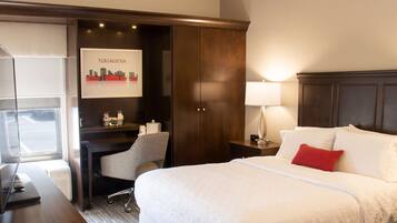 A hotel room with a bed, a desk, a chair, a nightstand, a lamp, and a framed picture of Tuscaloosa.
