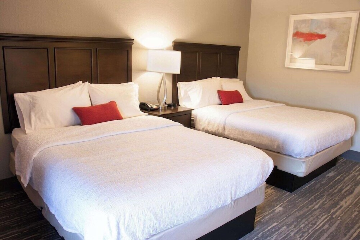 A hotel room with two beds, each with white bedding and red pillows, a nightstand with a lamp, and a framed abstract painting on the wall.
