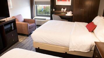 A hotel room with a large bed, a smaller bed, a desk with a chair, a TV, and a window with a cityscape view.