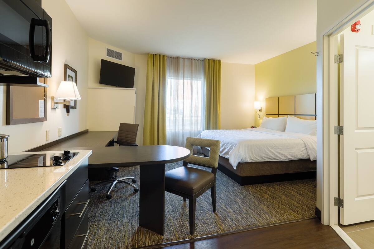 Een hotelkamer met een bed, een bureau met stoel, een magnetron, een televisie en een raam met gordijnen.