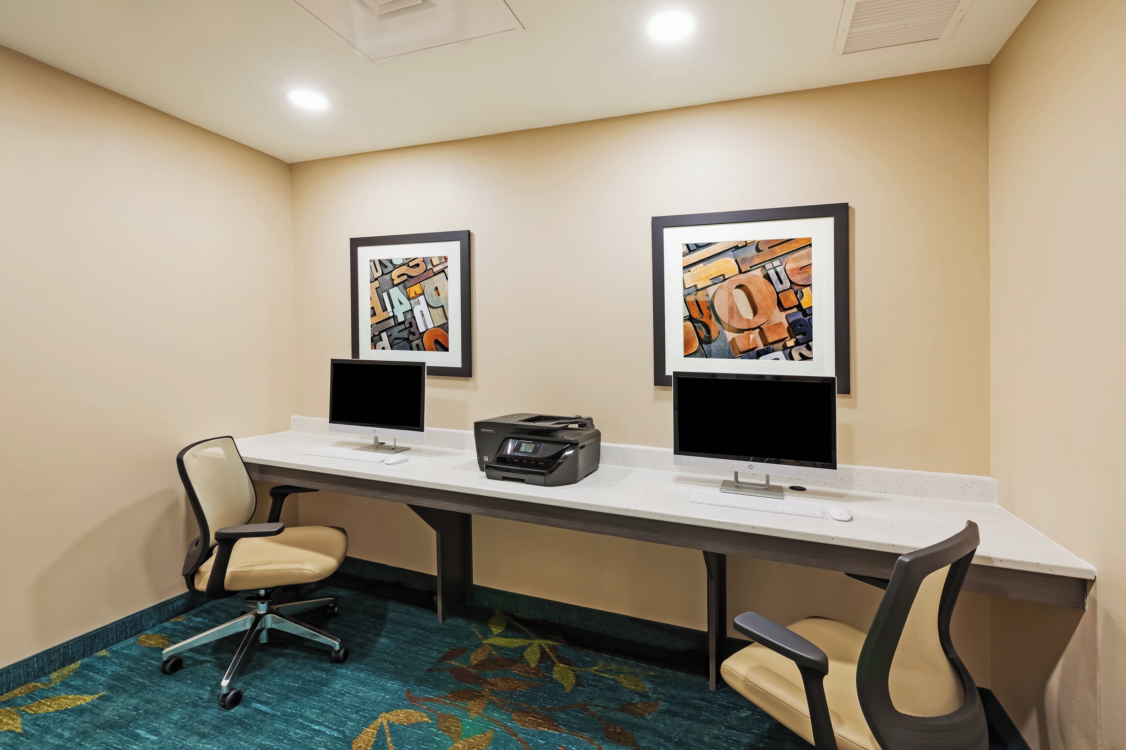 A conference room with two desks, each with a computer monitor, keyboard, and mouse. There are two framed artworks on the wall above the desks.