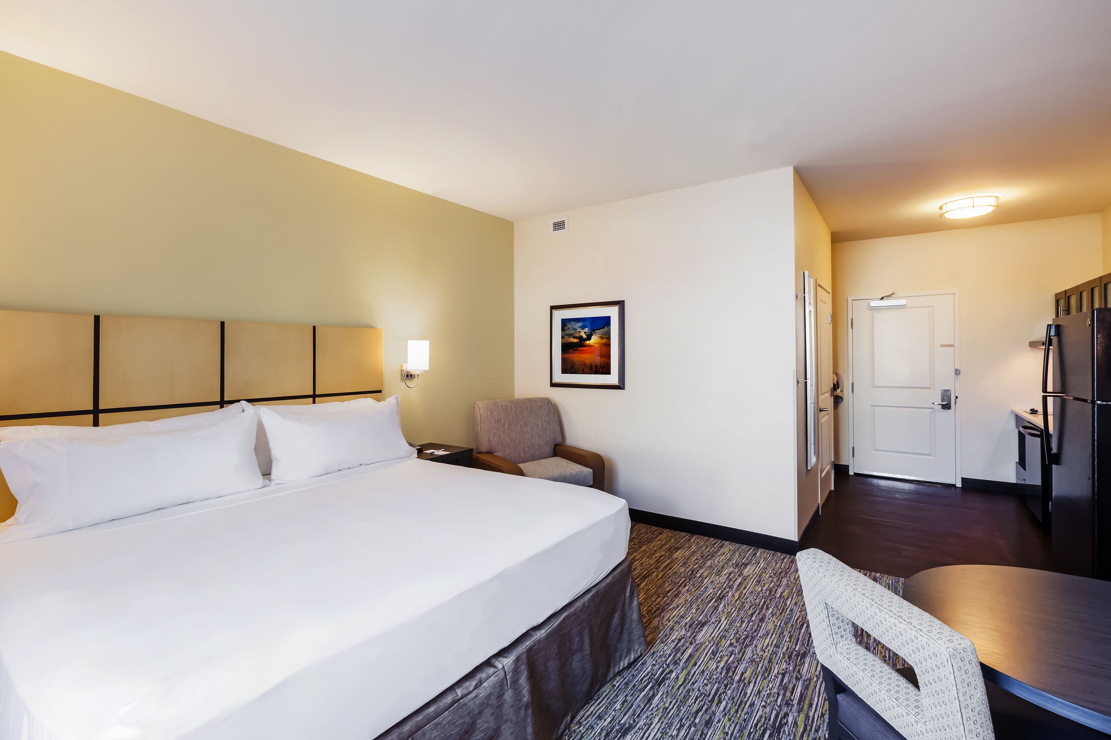 Habitación de hotel con una cama grande, un escritorio, una silla, una mesita, un cuadro en la pared y una puerta que da a la cocina.