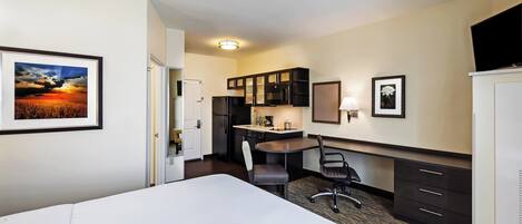 A hotel room with a bed, desk, chair, kitchenette, and a framed picture on the wall.