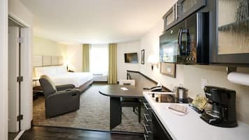 A hotel room with a bed, a sofa, a dining table, a kitchen area with a microwave, a coffee maker, and a TV.